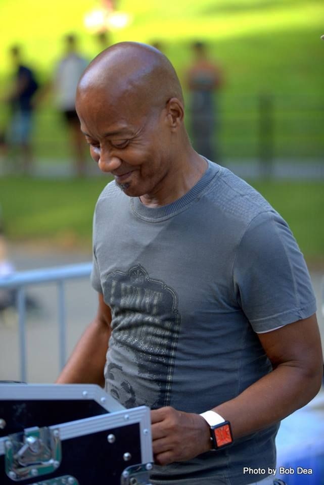 Man smiling while looking down, standing outdoors with blurred background.