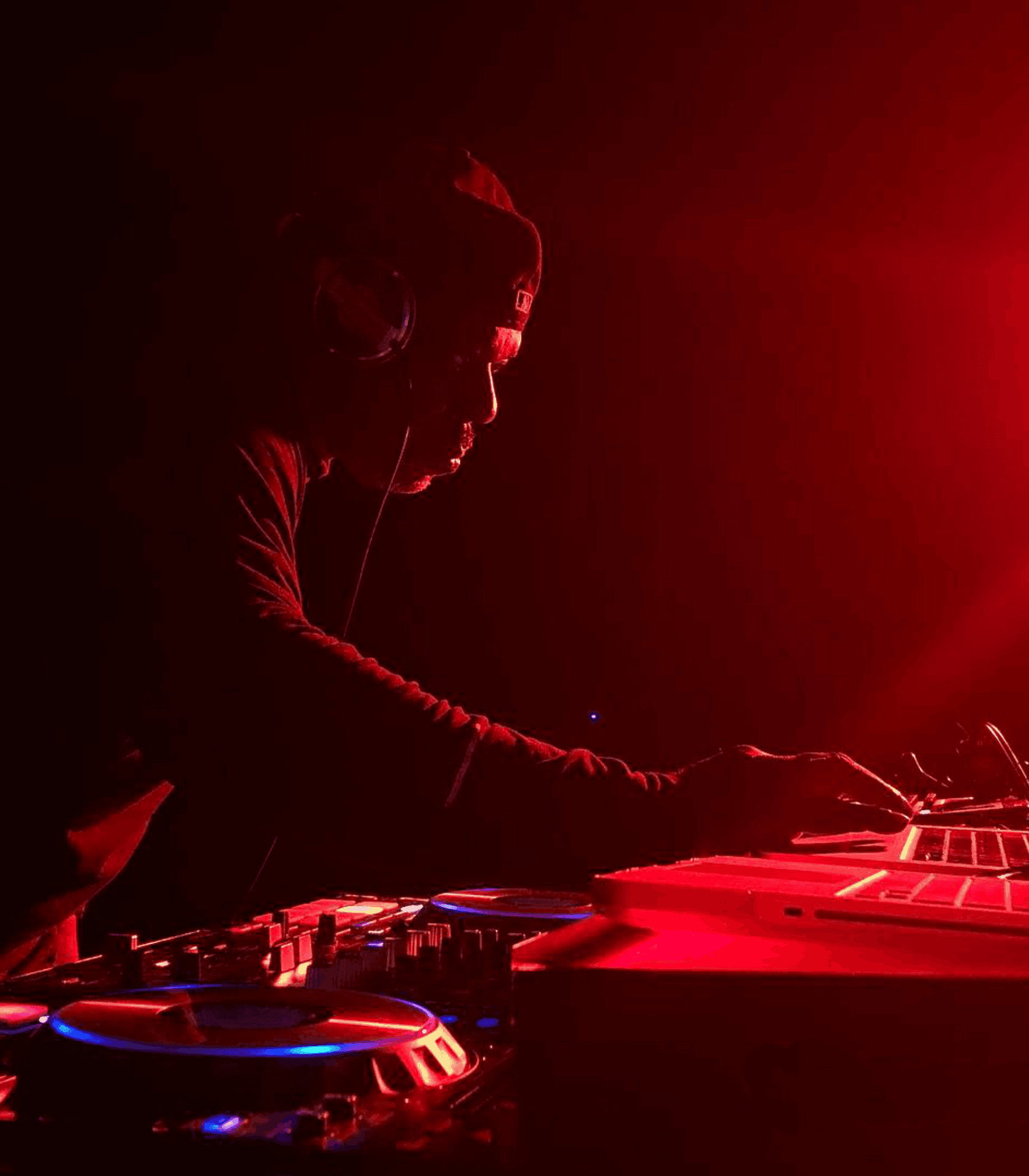 DJ in a dark room with red lighting, wearing headphones and operating DJ equipment.