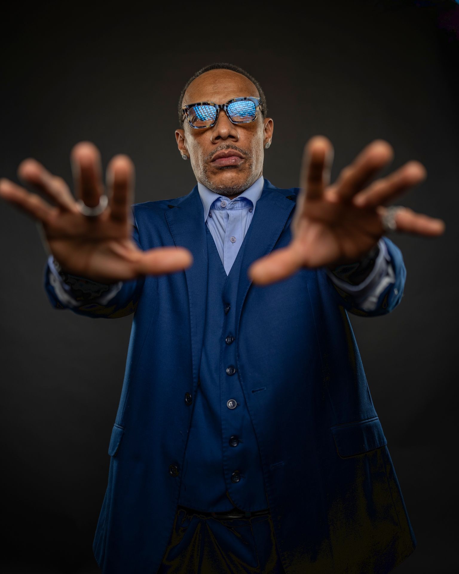 Man in blue suit with reflective sunglasses reaching forward against a dark background.